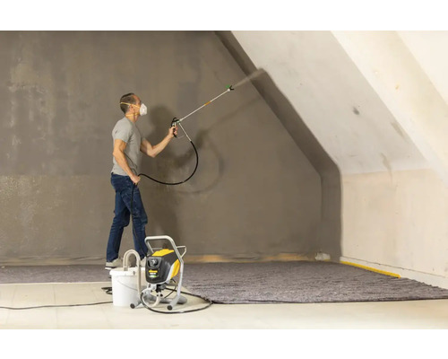 Muž strieka farbu pomocou striekacej pištole na stenu v miestnosti.