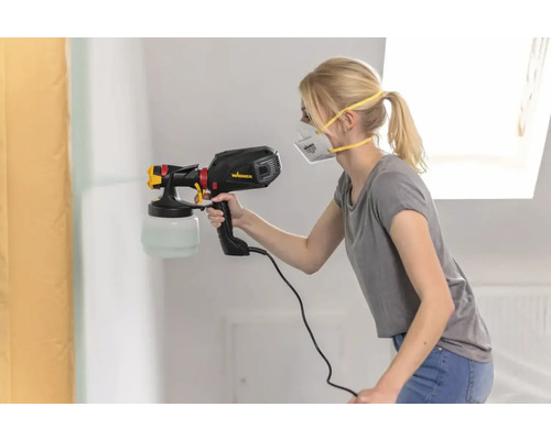 Žena strieka farbu na stenu pomocou striekacej pištole.
