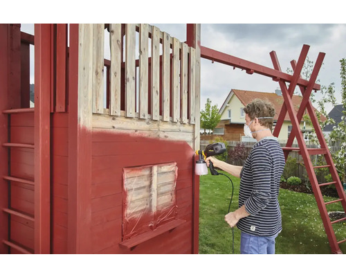 Osoba strieka drevený domček na hranie pomocou striekacej pištole na farbu