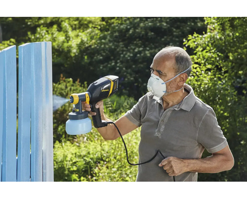Osoba používa striekaciu pištoľ na natieranie dreveného plota. Osoba má na sebe ochrannú masku.