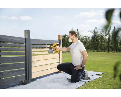 Muž natiera drevený plot striekacou pištoľou na farbu v záhrade.