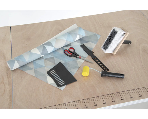 Tapeta s geometrickým vzorom, štetka na lepidlo, prítlačný valček, tapetovací nôž, prítlačná špachtľa a nožnice na stole.