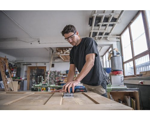 Muž brúsi drevené dvere akumulátorovou vibračnou brúskou v dielni.