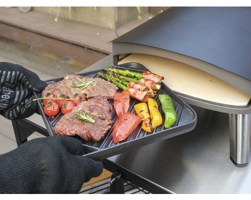 Osoba v kuchynských rukaviciach drží grilovaciu tácku s grilovaným steakom, špargľou, paradajkami a paprikami pred pecou na pizzu.