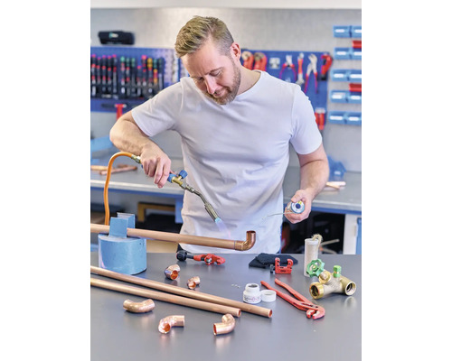 Muž spájkuje medenú rúrku spájkovačkou. Na stole ležia medené rúrky, tvarovky a náradie.