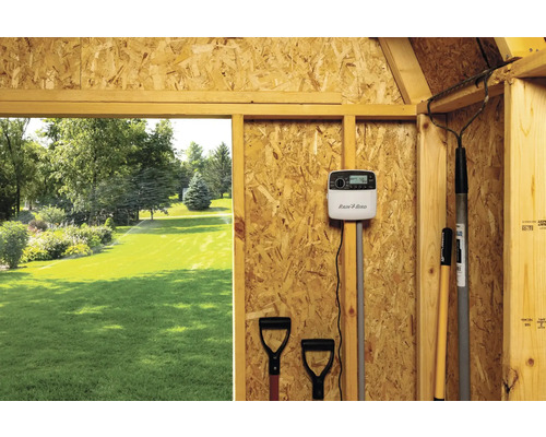 Scéna záhradného zavlažovacieho systému s ovládačom Rain Bird a záhradným náradím v drevenej kôlni, výhľad na trávnik s aktívnym zavlažovaním