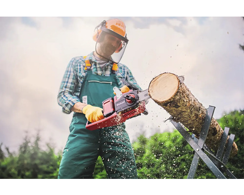 Muž s ochranným odevom reže motorovou pílou Einhell kmeň stromu na kozlíku