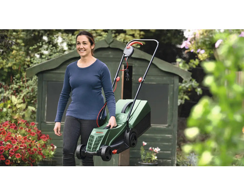 Žena nesie elektrickú kosačku na trávu v záhrade pred kôlňou na náradie.