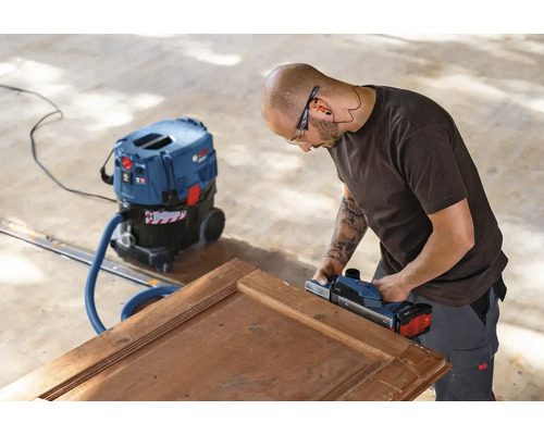 Remeselník hobľuje drevené dvere elektrickým hoblíkom a vysávačom