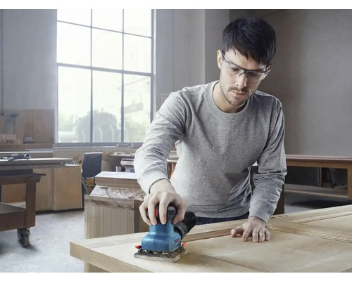 Muž brúsi drevo vibračnou brúskou v dielni.