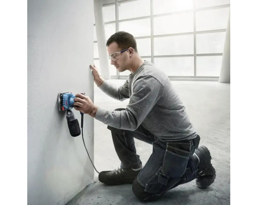 Muž brúsi stenu elektrickou brúskou vo svetlej miestnosti.