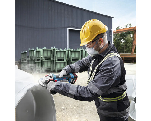 Muž s ochrannou prilbou, ochrannými okuliarmi a respirátorom brúsi obrobok pomocou akumulátorovej uhlovej brúsky.