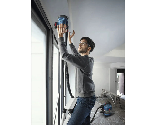 Muž na rebríku brúsi strop stropnou brúskou, pripojený k vysávaču