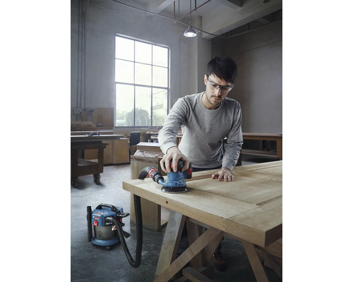 Muž brúsi drevené dvere pomocou excentrickej brúsky a vysávača v dielni.