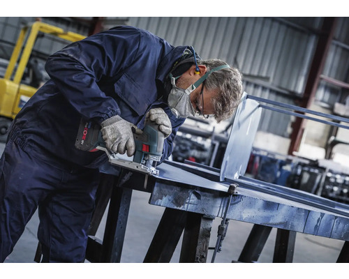Muž s ochrannou maskou, rukavicami a chráničmi sluchu reže kus kovu akumulátorovou priamočiarou pílou