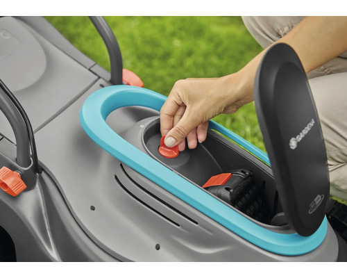 Ruka vkladá červené tlačidlo do kosačky na trávu s otvorenou priehradkou na batérie. Je viditeľné logo Gardena.