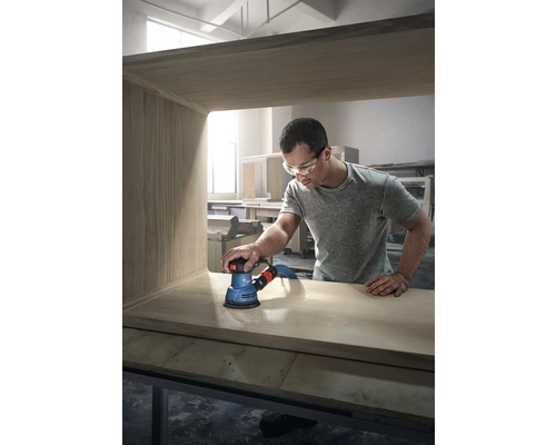 Muž brúsi drevený kus nábytku excentrickou brúskou značky Bosch.