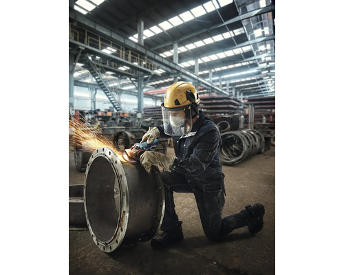 Robotník brúsi brúskou rúru v továrnej hale a má na sebe ochranný odev.