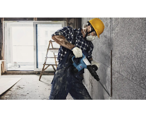 Stavebný robotník používa zbíjacie kladivo na betónovej stene vo svetlej miestnosti.