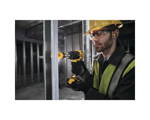 Muž s ochrannou prilbou, okuliarmi a ochranou sluchu vŕta žltým akumulátorovým vŕtacím skrutkovačom otvor do steny.