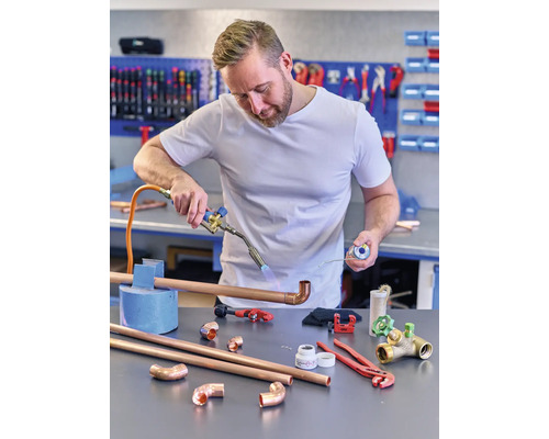 Inštalatér spájkuje medené rúry horákom, zatiaľ čo na ponku ležia rôzne nástroje a rúrkové diely.