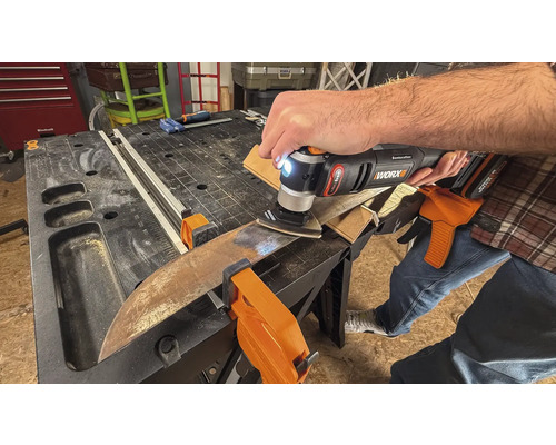 Osoba používa multifunkčný nástroj na brúsenie obrobku, ktorý je pripevnený svorkami k pracovnému stolu.