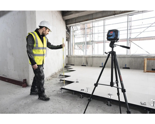 Remeselník s laserovou vodováhou a nivelačnou latou pri inštalácii zdvojenej podlahy.