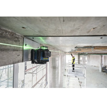 Zelený čiarový laser je pripevnený k betónovému nosníku a premieta zelenú laserovú čiaru. V pozadí stojí pracovník na lešení.