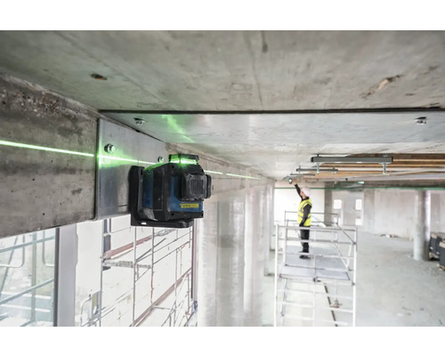 Zelený čiarový laser je pripevnený k betónovému nosníku a premieta zelenú laserovú čiaru. V pozadí stojí pracovník na lešení.