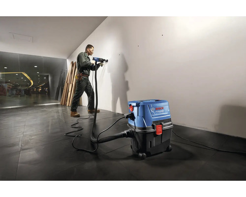 Remeselník vŕta vŕtačkou do steny, zatiaľ čo priemyselný vysávač odsáva prach.