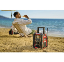 Stavebné rádio Einhell na pláži s osobou v pozadí, ktorá si vychutnáva pitie.
