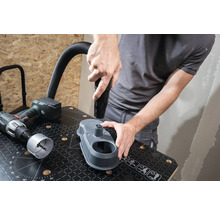 Remeselník pripevňuje hubicu vysávača k odsávaniu prachu z vŕtania na pracovnom stole. Vedľa leží vŕtačka a dierovacia pílový list.