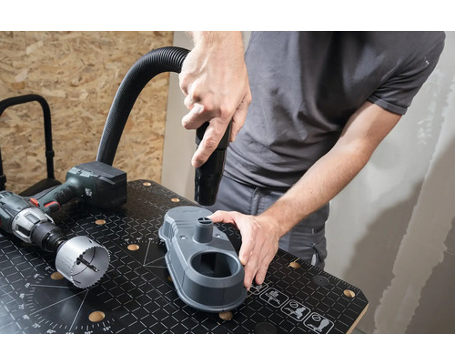 Remeselník pripevňuje hubicu vysávača k odsávaniu prachu z vŕtania na pracovnom stole. Vedľa leží vŕtačka a dierovacia pílový list.