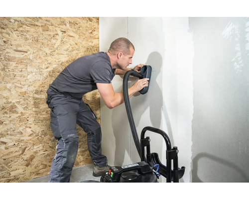 Muž brúsi stenu pomocou brúsky a vysávača
