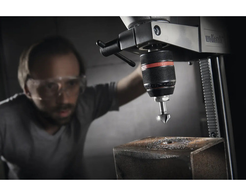 Stojanová vŕtačka s mužom s ochrannými okuliarmi, ktorý vŕta otvor do kusu kovu.