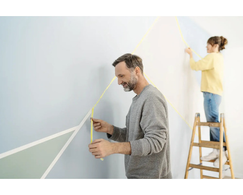 Dve osoby zdobia stenu maskovacou páskou a geometrickými tvarmi.