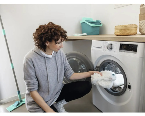 Žena vkladá biely návlek na mop do práčky vo svetlej miestnosti. Je tu tiež vidieť čistiace vedro a mop.