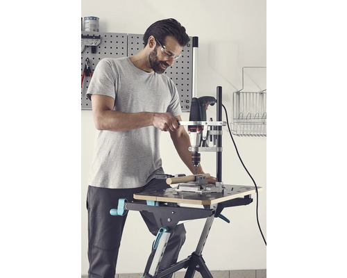 Muž s ochrannými okuliarmi vŕta vŕtačkou do obrobku na pracovnom stole.