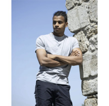 Mladý muž so zapletenými vlasmi sa opiera o kamenný múr so založenými rukami, má na sebe tričko a kapsáčové nohavice.