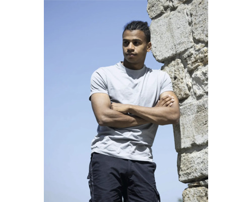 Muž so skríženými rukami, oblečený v tričku a šortkách, stojí pri kamennej stene