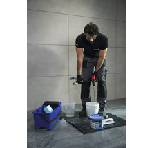 Remeselník mieša lepidlo na dlaždice s miešadlom farieb pred obloženou stenou. Modrý čistiaci vedro a nádoba s lepidlom sú pripravené.