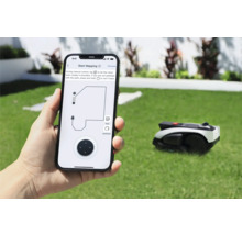 Osoba drží smartfón s aplikáciou na ovládanie robotickej kosačky na trávu. V pozadí je robotická kosačka na trávniku.