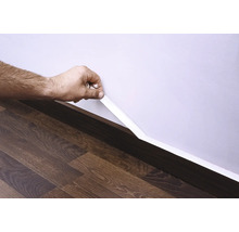 Osoba pripevňuje bielu podlahovú lištu k stene nad tmavou drevenou podlahou.