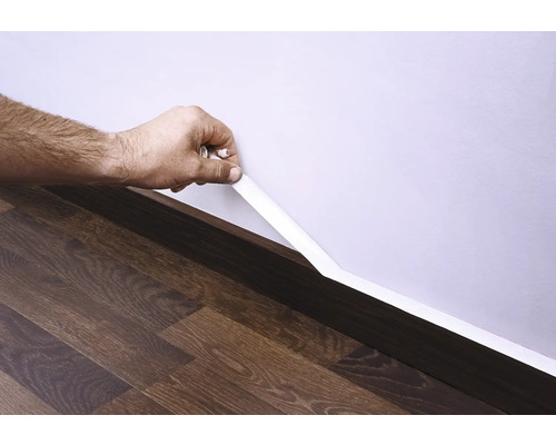 Upevnenie bielej podlahovej lišty na stenu nad tmavou drevenou podlahou.