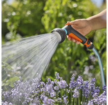 Ruka drží záhradnú hadicu so sprchovou hlavicou, ktorá strieka vodu na kvety.