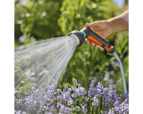 Ruka drží záhradnú hadicu so sprchovou hlavicou, ktorá strieka vodu na kvety.