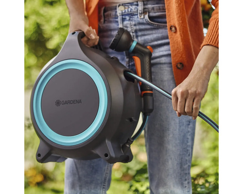 Osoba drží hadicový box Gardena so záhradnou hadicou a postrekovačom.