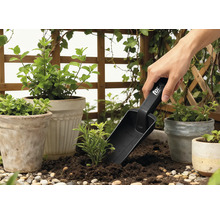 Záhradná scéna s kvetináčmi, okolitou zeleňou a osobou používajúcou sadzačku.