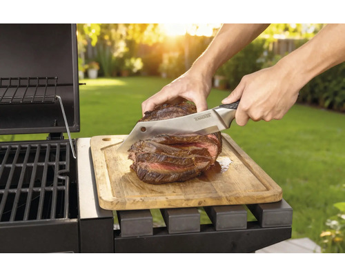 Pripravený steak sa krája nožom na drevenej doske vedľa grilu.