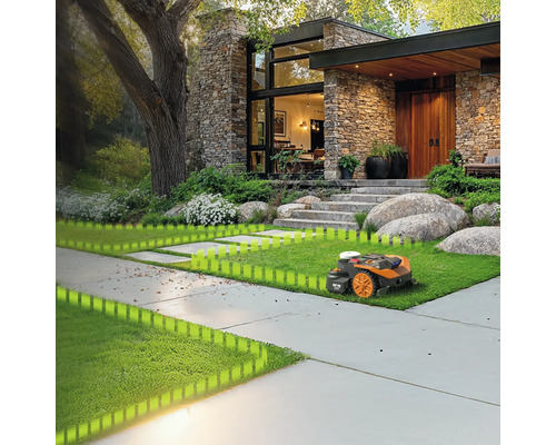 Robotická kosačka na trávniku pred domom s kamennou fasádou a veľkým stromom.